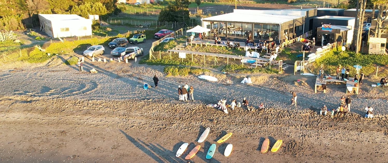 Vista aérea del complejo Cura-Quillas a la orilla de Playa Curaquilla: la sala y el café frente al mar, tablas de surf sobre la arena y el estacionamiento en el acceso